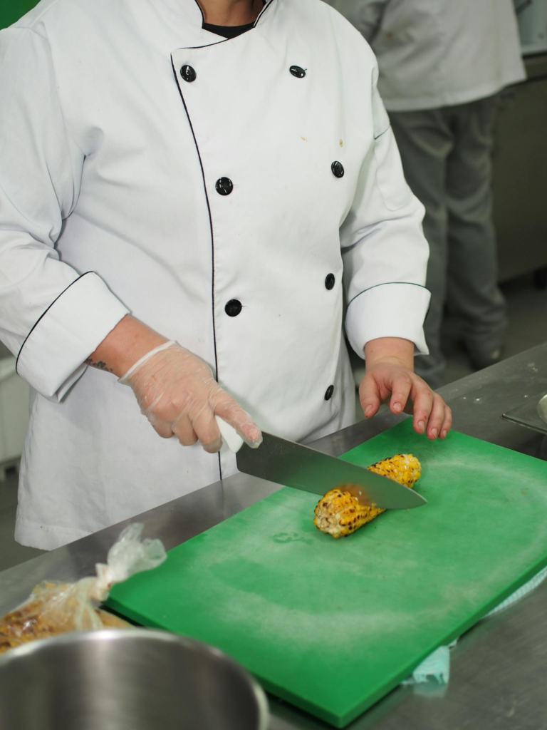 Chef in white uniform slicing corn on green cutting board in professional kitchen setting.