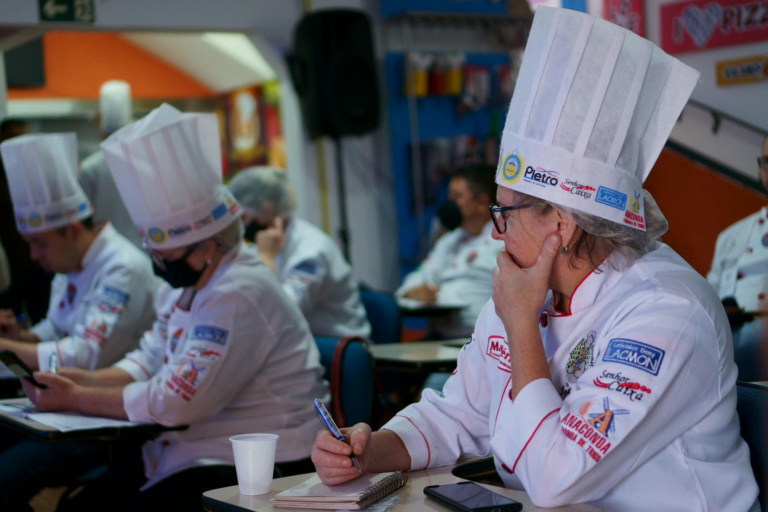 Group of chefs in a culinary class, attentively taking notes while wearing traditional hats and uniforms.