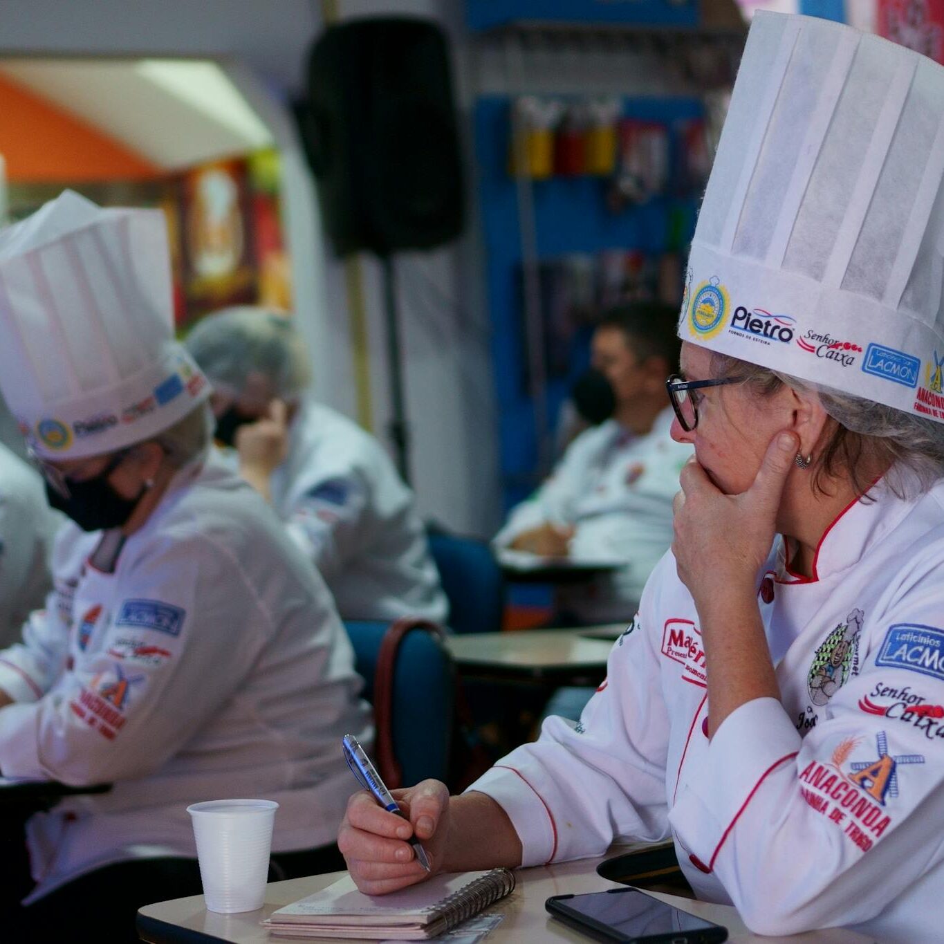 Group of chefs in a culinary class, attentively taking notes while wearing traditional hats and uniforms.