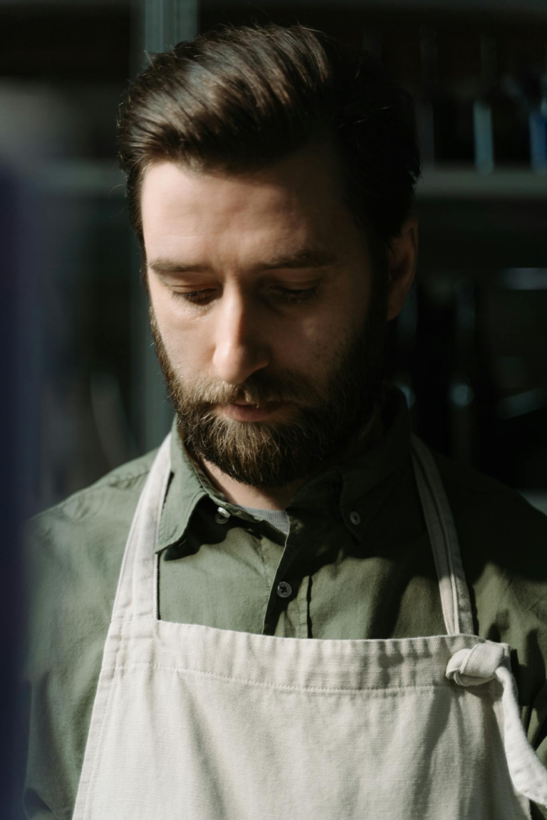 Young male chef in a green shirt and apron, looking down thoughtfully in a kitchen environment.